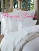Pioneer Linens Catalog: Luxury white bedding with intricate embroidery and plush pillows on a wooden bed frame.